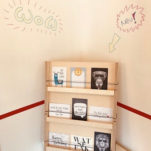 Puede incluir: Un soporte de madera con tres estantes, cada uno con una variedad de tarjetas de felicitaci&oacute;n. Las tarjetas presentan diferentes dise&ntilde;os, incluyendo una llama, un mono, un le&oacute;n y una tarjeta de texto que dice "Sopa del d&iacute;a". El soporte est&aacute; contra una pared blanca con rayas rojas y blancas en la parte inferior.