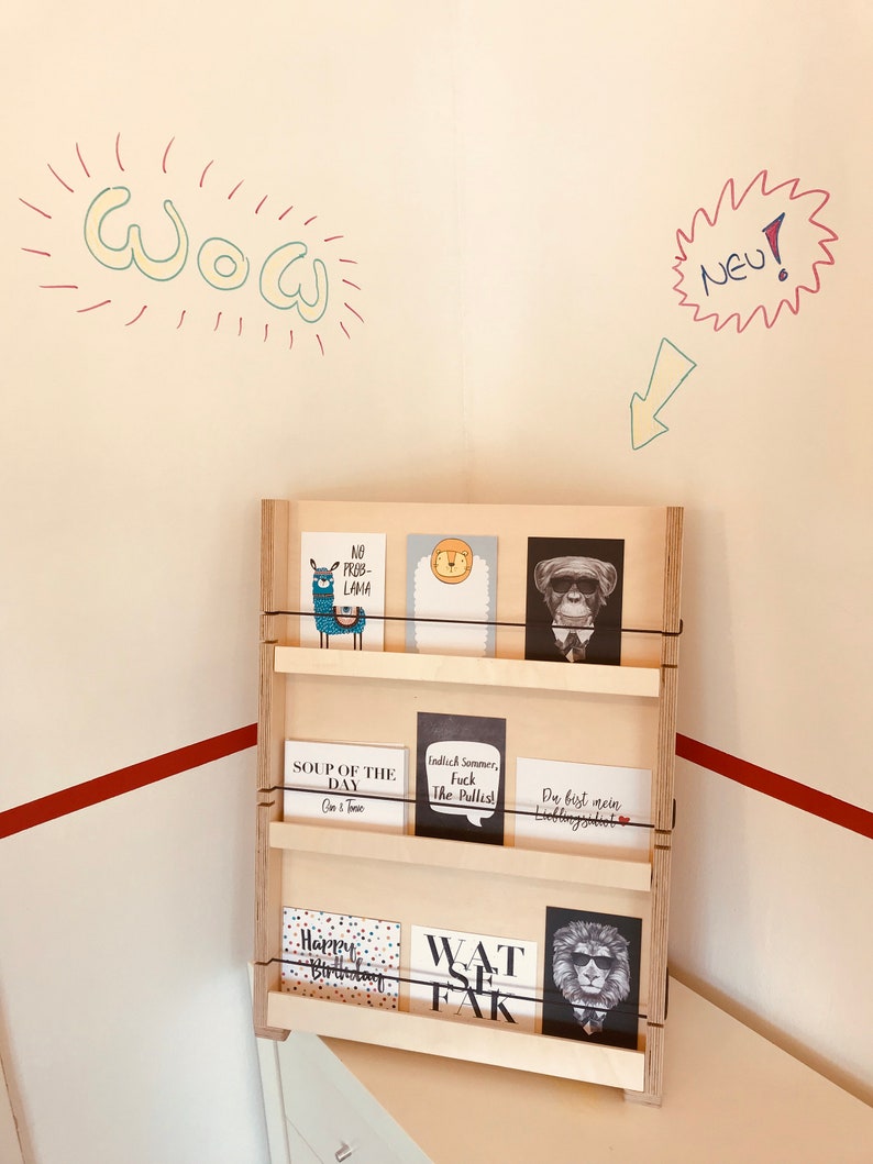 Puede incluir: Un soporte de madera con tres estantes, cada uno con una variedad de tarjetas de felicitaci&oacute;n. Las tarjetas presentan diferentes dise&ntilde;os, incluyendo una llama, un mono, un le&oacute;n y una tarjeta de texto que dice "Sopa del d&iacute;a". El soporte est&aacute; contra una pared blanca con rayas rojas y blancas en la parte inferior.