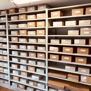 May include: A storage room with multiple metal shelves filled with various sized cardboard boxes. The boxes are primarily tan and light gray, with some labeled. The shelves are attached to the wall and are evenly spaced, creating a grid-like pattern.