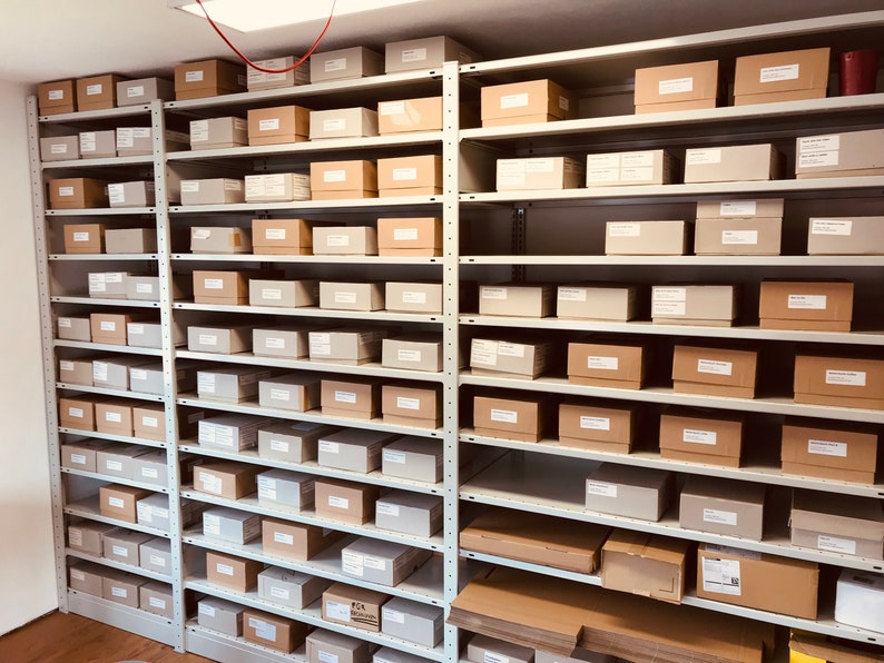 May include: A storage room with multiple metal shelves filled with various sized cardboard boxes. The boxes are primarily tan and light gray, with some labeled. The shelves are attached to the wall and are evenly spaced, creating a grid-like pattern.