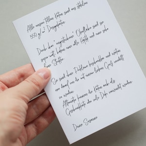 May include: A white card with handwritten black text is held by a hand. The card's text is in German, with a signature at the bottom. The card appears to be a note or a message, possibly for a gift or special occasion.