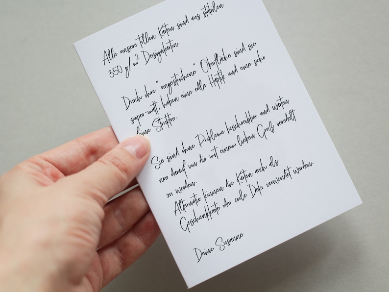 May include: A white card with handwritten black text is held by a hand. The card's text is in German, with a signature at the bottom. The card appears to be a note or a message, possibly for a gift or special occasion.