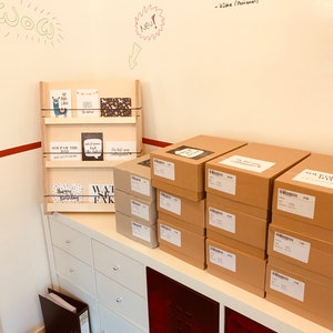 May include: A wooden display shelf holding various greeting cards with different designs and text. Several cardboard boxes are stacked on a white cabinet. The wall behind the shelf has handwritten text and drawings.