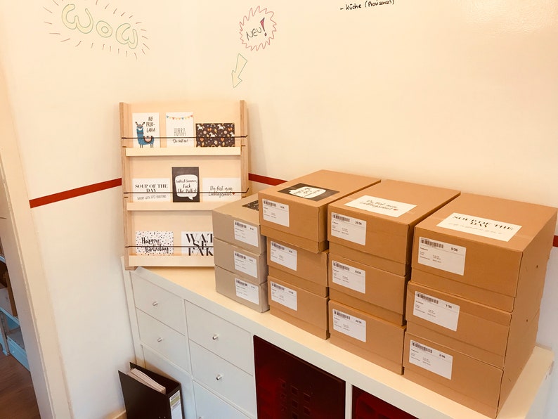 May include: A wooden display shelf holding various greeting cards with different designs and text. Several cardboard boxes are stacked on a white cabinet. The wall behind the shelf has handwritten text and drawings.