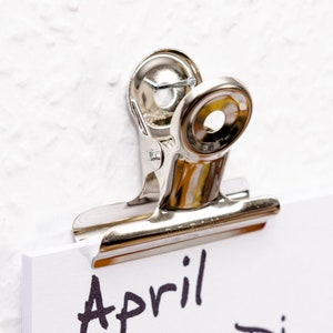 Könnte beinhalten: Ein silberner Metall-Binderclip hält ein weißes Blatt Papier mit dem Wort "April" in schwarzer Tinte.