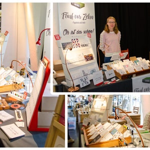 May include: A display of greeting cards and stationery at a craft fair. The booth features various card designs, displayed in wooden trays and on angled display stands. The booth's sign reads "F&uuml;nf vor Zehn". A woman stands behind the table.