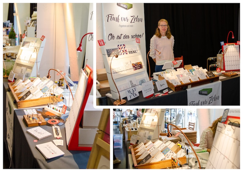 May include: A display of greeting cards and stationery at a craft fair. The booth features various card designs, displayed in wooden trays and on angled display stands. The booth's sign reads "F&uuml;nf vor Zehn". A woman stands behind the table.