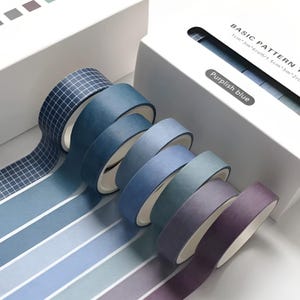 Może przedstawiać: Kolekcja rolek taśmy washi w odcieniach niebieskiego i fioletowego. Jedna rolka ma wzór kratki, inne są gładkie lub w paski. Na opakowaniu widnieje napis "BASIC PATTERN TAPE" i "Purplish blue".