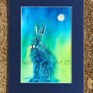 May include: Watercolor painting of a blue hare under a full moon, set against a gradient of green and blue. The artwork is matted in white and framed in a dark blue mat. The artist's signature is visible.