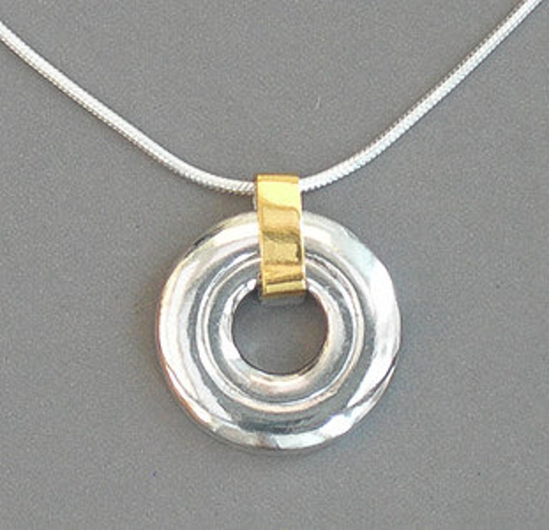 May include: Silver pendant necklace with a gold band around the center of the circle. The pendant has a textured, concentric circle design.