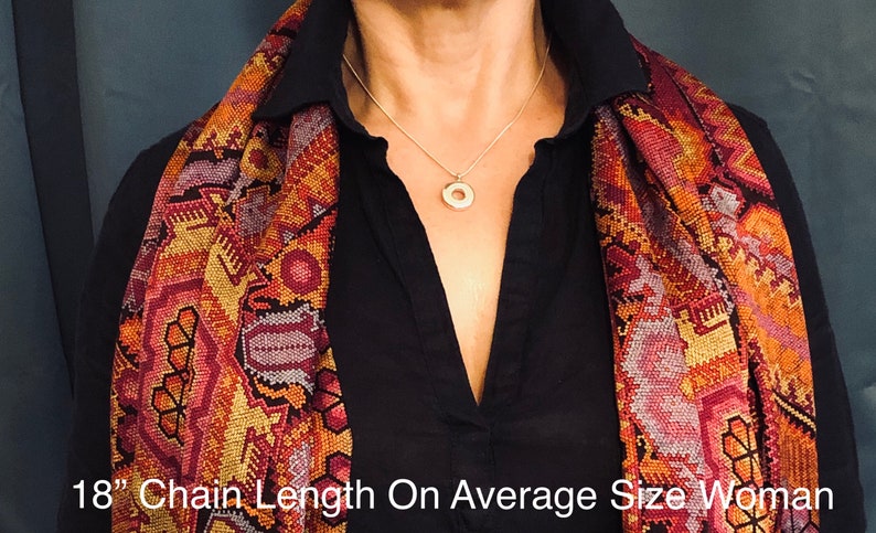 May include: A person wearing a black shirt with a colorful patterned scarf. The person is wearing a silver chain necklace with a round pendant. The text "18" Chain Length On Average Size Woman" is at the bottom of the image.