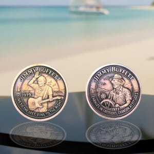 May include: Two bronze-colored commemorative coins featuring Jimmy Buffett. One coin shows Buffett playing guitar with the text "FINS TO THE LEFT, FINS TO THE RIGHT" and the year 1979. The other coin shows Buffett at the helm with the text "CHANGES IN LATITUDES, CHANGEN ATTITUDES" and the year 1977.