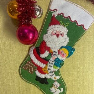 May include: A green felt Christmas stocking with a white felt cuff. The stocking features a Santa Claus figure, a child holding a toy, and a white cat. The stocking has a gold trim and a bell at the bottom. The stocking has a list of toys written on it: "Dog Game, Car, Bike, Top, Ball, Yo-Yo, Cap."
