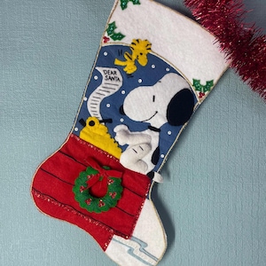 May include: A Christmas stocking featuring Snoopy and Woodstock. The stocking has a red base with a green wreath, a blue section with Snoopy, and a white top with holly accents. The words "DEAR SANTA" are visible on a scroll.