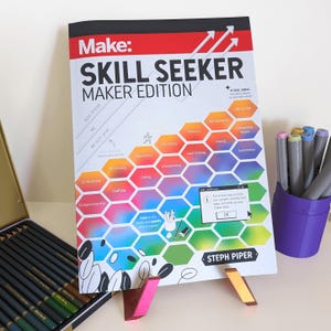 Può includere: Un libro colorato intitolato "Make: Skill Seeker Maker Edition" di Steph Piper. La copertina presenta un motivo a nido d'ape con diverse abilità elencate in ogni esagono, come la codifica, la falegnameria e la robotica. Il libro è su un supporto con un portapenne viola e matite colorate.