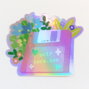 Könnte beinhalten: Ein holografischer Aufkleber mit einer Diskette mit dem Text "self care.exe" und einem Blumenmuster. Die Diskette ist rosa und lila mit einem blauen Etikett, auf dem "1.44 mb" steht.