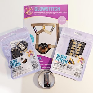 May include: A GlowStitch craft kit featuring a purple instructional pamphlet, two sealed plastic pouches containing battery holders and LED strips, and two AAA batteries placed inside a small circular roll of conductive tape.