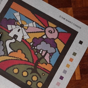 Puede incluir: Un lienzo de punto de aguja colorido con un diseño caprichoso que presenta un molino de viento, un sol y una nube. El lienzo está rodeado por un borde negro e incluye una clave de color para los diferentes hilos utilizados en el diseño. El texto "@THE NAKED CANVAS" está impreso en la esquina superior derecha.
