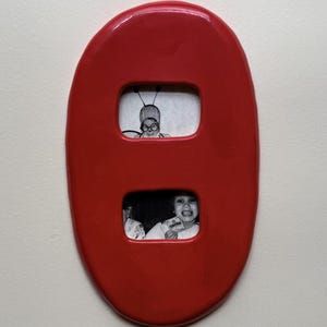 Pode incluir: Uma moldura de parede oval de cerâmica vermelha brilhante com duas aberturas retangulares. A abertura superior exibe uma fotografia a preto e branco de uma pessoa com óculos e antenas, enquanto a inferior mostra uma foto a preto e branco de uma pessoa sorridente.