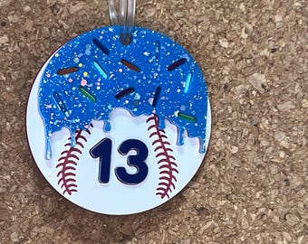 May include: A round baseball-themed ornament with a blue, glittery top resembling a donut with sprinkles. The number 13 is in the center, with red baseball stitching. A clear ribbon is attached for hanging.