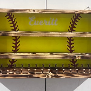 Puede incluir: Estante de exhibición de softbol de madera con fondo amarillo y diseño de costuras marrones. El nombre "Everitt" está escrito en blanco. Cuenta con múltiples estantes y clavijas para exhibir artículos.