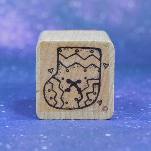 Puede incluir: Un sello de goma cuadrado montado en madera con una ilustración en tinta negra de un calcetín navideño. El diseño incluye líneas en zigzag, puntos pequeños, un detalle de punto de cruz central, tres corazones pequeños y un símbolo de copyright.