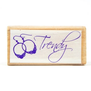 May include: A wooden stamp with the word "Trendy" in cursive script and an illustration of hoop earrings. The earrings are purple and have a decorative design. The stamp is on a white background.