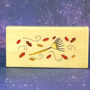 May include: A wooden stamp with a design of a rake and leaves blowing in the wind. The rake is yellow and the leaves are red and orange.