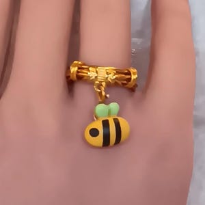 May include: A gold ring with a dangling bee charm. The bee is yellow with black stripes and green wings.