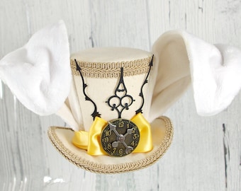 May include: A cream-colored top hat featuring two large, plush white rabbit ears extending from the sides. The hat is decorated with a gold braided trim, a black filigree clock hand ornament, and a bright yellow ribbon bow with a clock face.