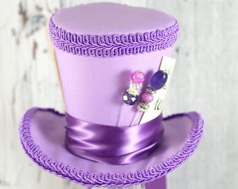 Pode incluir: Um chapéu alto lilás com aba larga e uma fita de cetim. O chapéu é adornado com trança decorativa e adornado com enfeites de contas e um pequeno cartão com o número 10. O chapéu é exibido em um suporte de vidro transparente.