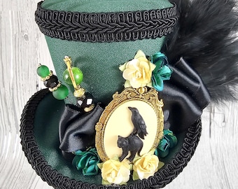 Puede incluir: Un sombrero de copa verde con ribete negro y una cinta negra. El sombrero está decorado con flores amarillas y verdes, un camafeo con marco dorado con un cuervo y un gato negro, y plumas negras. Un adorno de cuentas añade detalles.