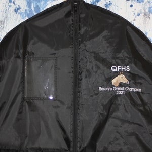Pu&ograve; includere: Borsa per abiti nera con chiusura a zip e una finestra trasparente in plastica. La borsa ha un logo ricamato che dice "QFHS Reserve Overall Champion 2021" con la testa di un cavallo.