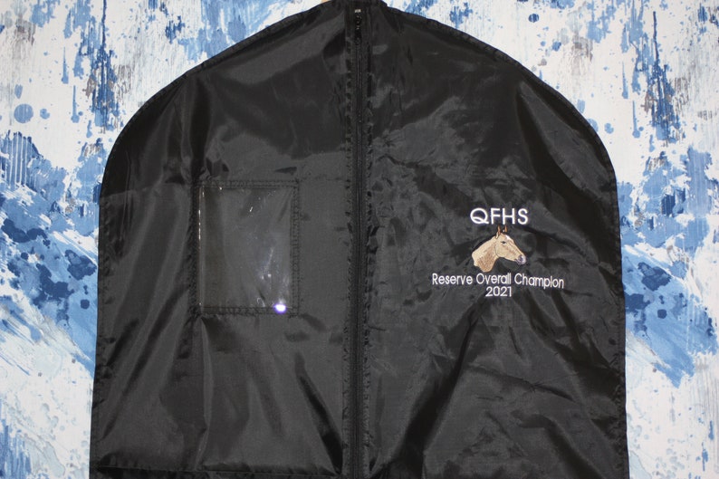 Pu&ograve; includere: Borsa per abiti nera con chiusura a zip e una finestra trasparente in plastica. La borsa ha un logo ricamato che dice "QFHS Reserve Overall Champion 2021" con la testa di un cavallo.