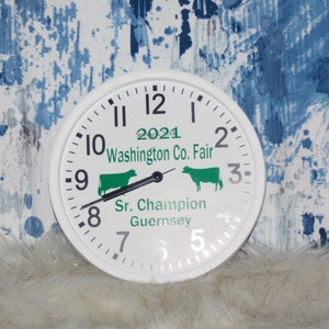 K&ouml;nnte beinhalten: Wei&szlig;e Uhr mit schwarzen Zeigern und Zahlen. Das Zifferblatt der Uhr tr&auml;gt den Text "2021 Washington Co. Fair" und "Sr. Champion Guernsey". Auf dem Zifferblatt befinden sich zwei gr&uuml;ne Kuhsilhouetten.