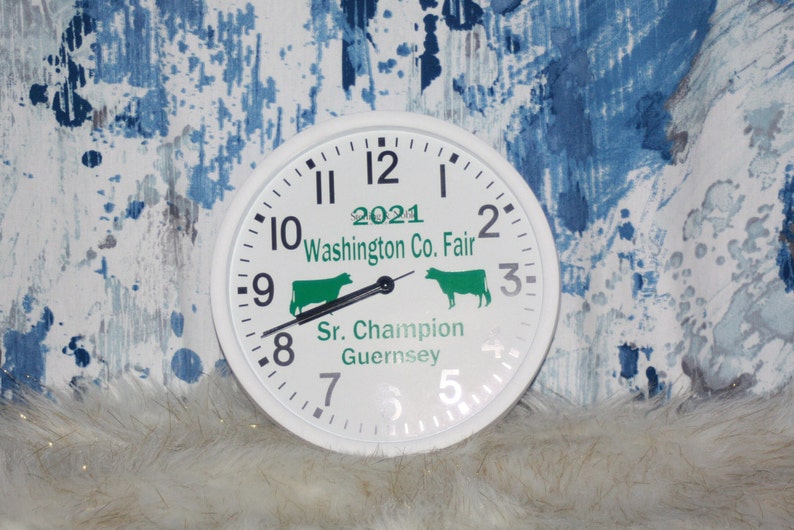 K&ouml;nnte beinhalten: Wei&szlig;e Uhr mit schwarzen Zeigern und Zahlen. Das Zifferblatt der Uhr tr&auml;gt den Text "2021 Washington Co. Fair" und "Sr. Champion Guernsey". Auf dem Zifferblatt befinden sich zwei gr&uuml;ne Kuhsilhouetten.