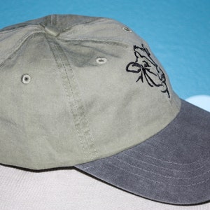 May include: A green and gray baseball cap with a black embroidered cow design on the front.