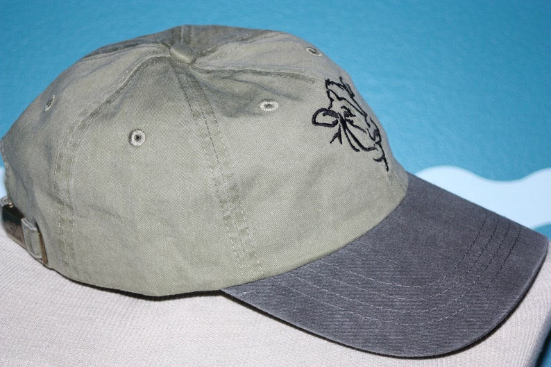 May include: A green and gray baseball cap with a black embroidered cow design on the front.
