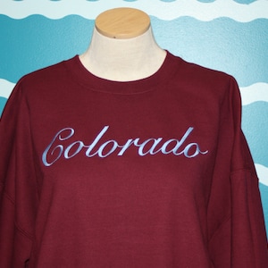 May include: A maroon sweatshirt with the word "Colorado" embroidered in light blue script across the chest. The sweatshirt is displayed on a mannequin with a wooden neck and shoulders.