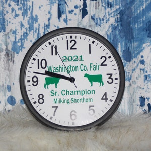 K&ouml;nnte beinhalten: Ein wei&szlig;es Zifferblatt mit schwarzen Zahlen und Zeigern. Das Zifferblatt hat eine gr&uuml;ne Kuhsilhouette und den Text "2021 Washington Co. Fair Sr. Champion Milking Shorthorn". Die Uhr hat einen schwarzen Rand.