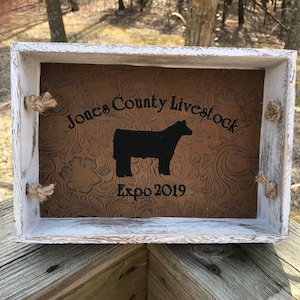 May include: A white wooden tray with rope handles. The tray has a brown leather-like lining with a black silhouette of a cow and the text "Jones County Livestock Expo 2019".