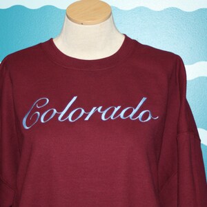 May include: A maroon sweatshirt with the word "Colorado" embroidered in light blue script across the chest. The sweatshirt is displayed on a mannequin with a neutral background.