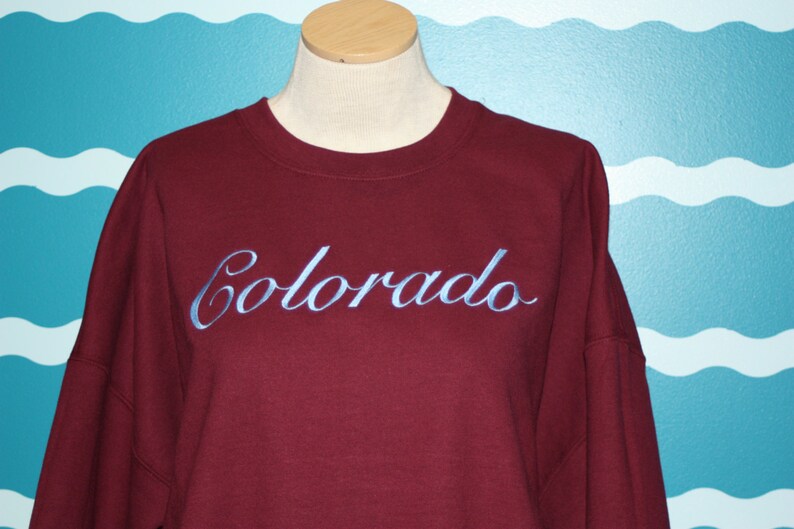 May include: A maroon sweatshirt with the word "Colorado" embroidered in light blue script across the chest. The sweatshirt is displayed on a mannequin with a neutral background.