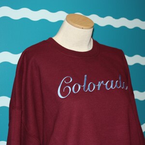 May include: A maroon sweatshirt with the word "Colorado" embroidered in a light blue script. The sweatshirt is displayed on a mannequin against a teal wall with white wave patterns.