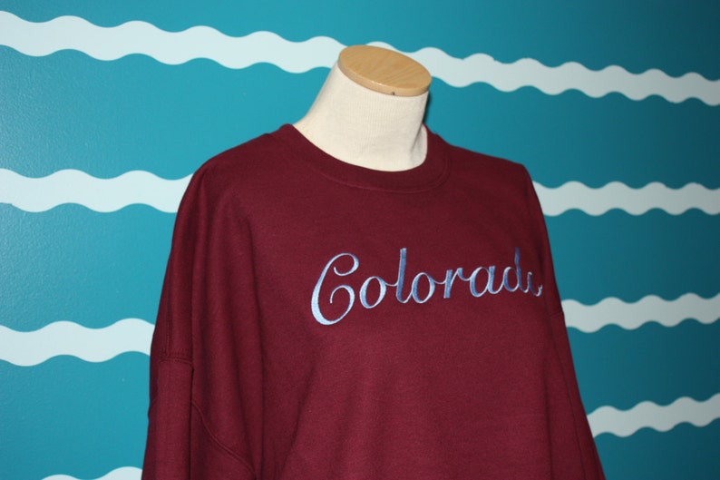 May include: A maroon sweatshirt with the word "Colorado" embroidered in a light blue script. The sweatshirt is displayed on a mannequin against a teal wall with white wave patterns.