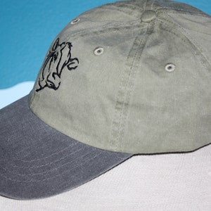 May include: A green and gray baseball cap with a black embroidered cartoon character on the front.