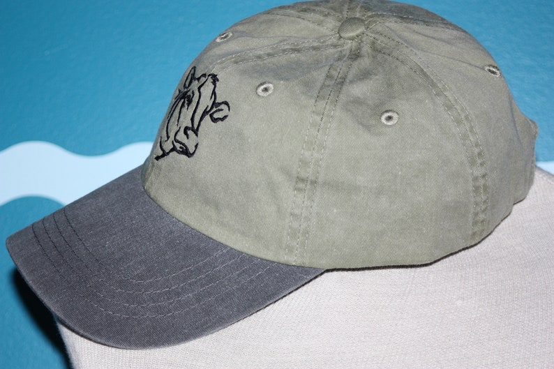 May include: A green and gray baseball cap with a black embroidered cartoon character on the front.