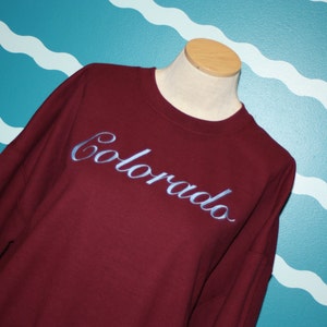 May include: A maroon sweatshirt with the word "Colorado" embroidered in a light blue script across the chest. The sweatshirt is displayed on a mannequin against a teal wall with white wave patterns.