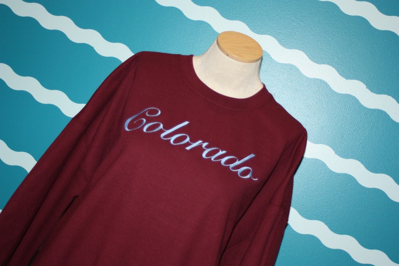 May include: A maroon sweatshirt with the word "Colorado" embroidered in a light blue script across the chest. The sweatshirt is displayed on a mannequin against a teal wall with white wave patterns.