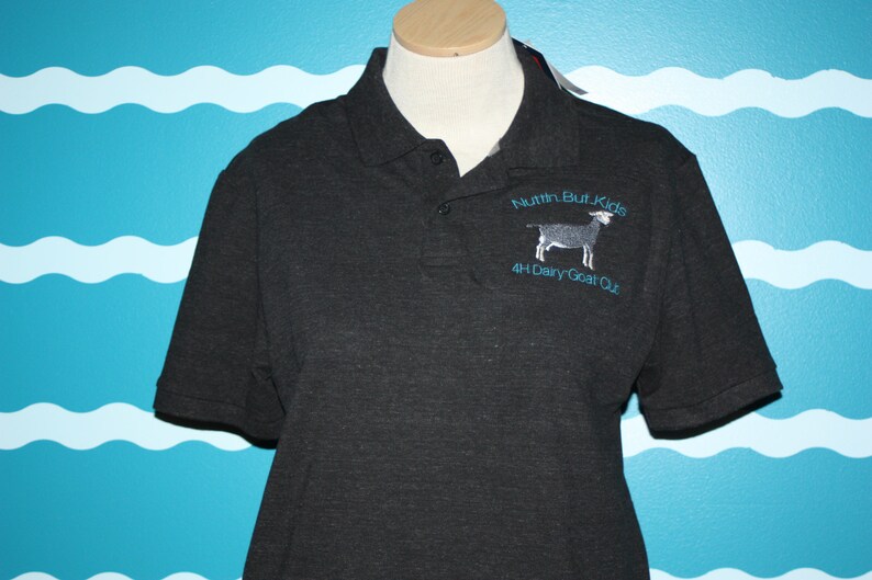 K&ouml;nnte beinhalten: Ein dunkelgraues Poloshirt mit einem blauen gestickten Logo, auf dem "Nuthin But Kids 4H Dairy Goat Club" steht und eine Ziege abgebildet ist.